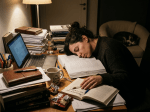 Tired student asleep on books at cluttered study desk with laptop and coffee cup